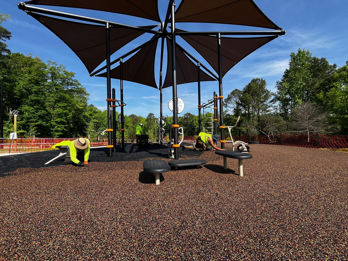 Workers installing playground equipment under large shaded canopies with colorful rubber flooring