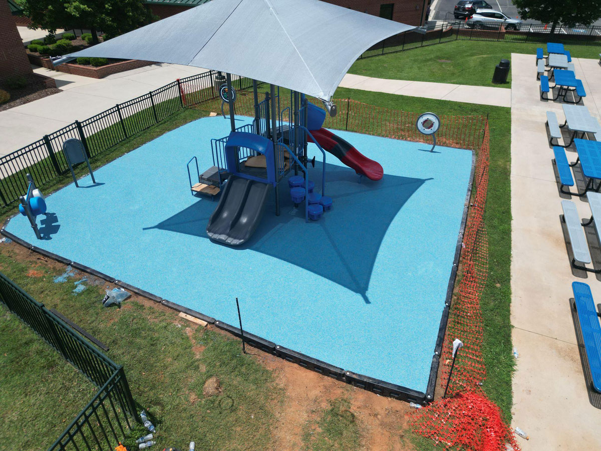 Playground with blue rubber flooring, slide, and shaded canopy in a fenced area